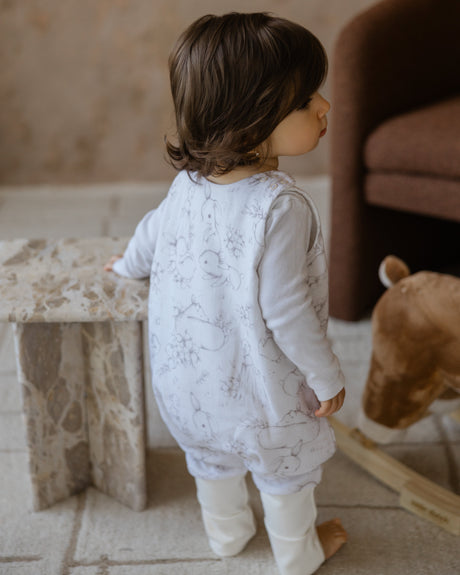 Child in a white onesie standing in a room with a teddy bear and a stone block.