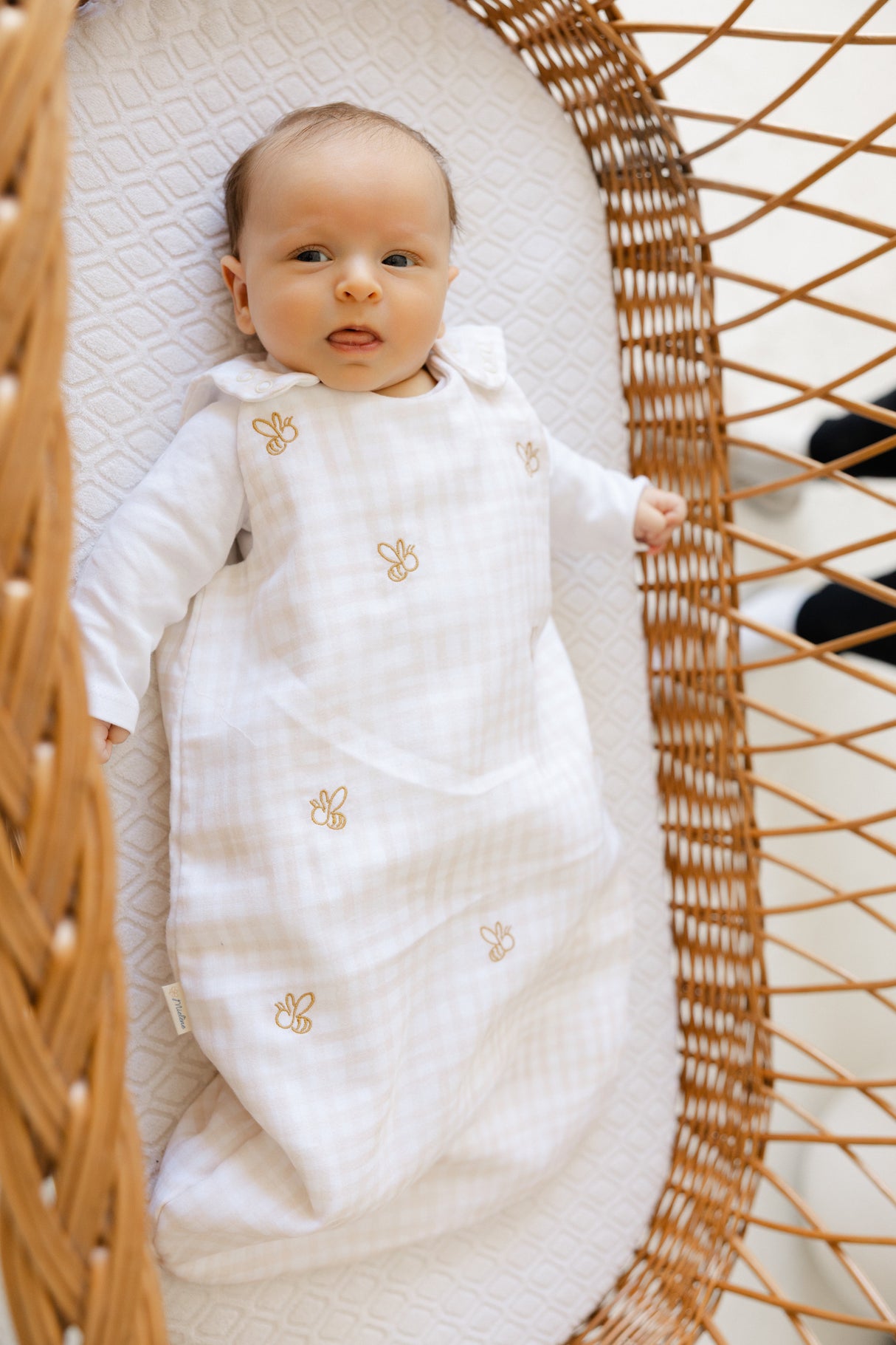 Baby in a white outfit with gold accents lying in a wicker crib.
