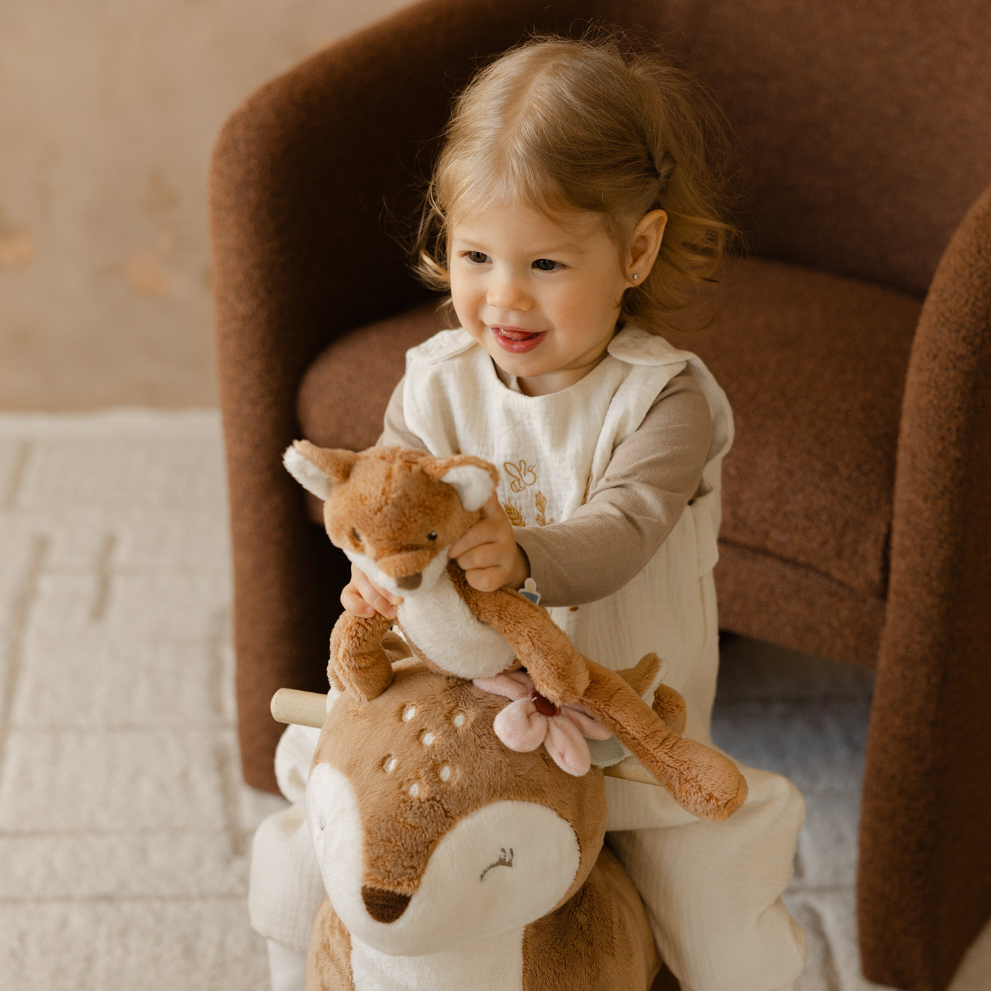 Child sitting on a plush deer toy chair holding a teddy bear in a cozy room.